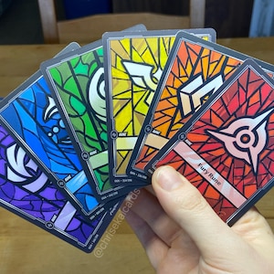 May include: A hand holds a fan of colorful cards with stained-glass style designs. Each card features a different color scheme, including purple, blue, green, yellow, and red. The cards have a black border and white text, with the words "Fury Rune" visible on one.