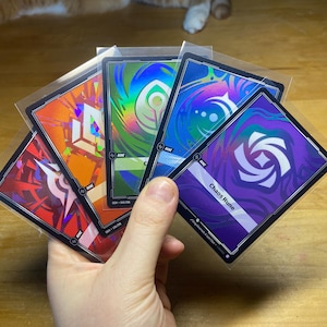 Puede incluir: Una mano sostiene un abanico de cinco cartas cuadradas en fundas protectoras. Cada carta presenta un diseño abstracto único en colores vibrantes como rojo, naranja, verde, azul y morado. Una carta tiene el texto "Chaos Rune".