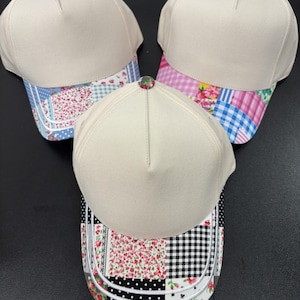 Könnte beinhalten: Drei Baseballkappen mit cremefarbenen Kronen und gemusterten Schirmen. Jede Kappe hat einen kleinen Knopf an der Oberseite, der zum Stoff des Schirms passt. Die Schirme zeigen Patchwork-Designs in Blau, Pink und Schwarz mit Blumen- und Karomustern.