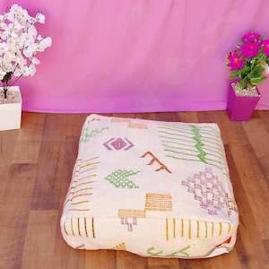 Puede incluir: Un puf cuadrado de color crema con un estampado geométrico en naranja, verde, morado y marrón. El cojín está sobre un suelo de madera, con flores rosas en una maceta blanca y flores rosas en una maceta rosa en el fondo.