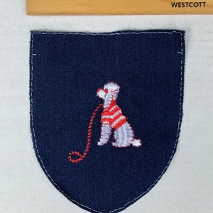 May include: A navy blue embroidered patch in a shield shape. The patch features a white dog with a red and white striped sweater and a red leash. A wooden ruler with inch markings is above the patch.