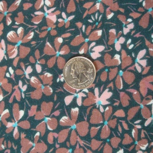 May include: Close-up of a fabric with a floral pattern. The design features small, stylized flowers in shades of brown and pink, set against a dark teal background. Each flower has a small, turquoise center. A U.S. quarter coin is included for scale.