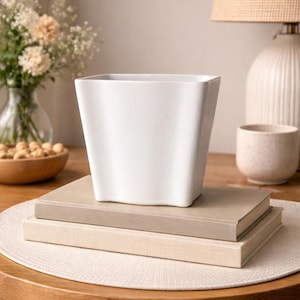 May include: A white square ceramic planter sits atop two books on a wooden table. The planter has a slightly tapered design. A vase of flowers and a lamp are in the background, creating a cozy, neutral-toned scene.