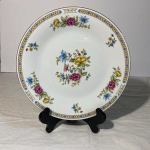 May include: White ceramic plate with a floral design. The plate features a gold rim and a border with blue and gold accents. The center of the plate has a cluster of flowers in pink, yellow, and blue. The plate is displayed on a black stand.
