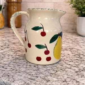 May include: Cream-colored ceramic pitcher with a handle, decorated with red cherries and a yellow pear. The pitcher has a green rim and is sitting on a speckled countertop. A decorative kitchen item.