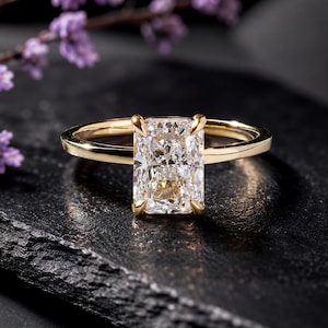 May include: A gold ring featuring a single rectangular radiant-cut diamond held by four prongs. The band is smooth and polished yellow gold, showcasing a clear, faceted gemstone that reflects light with a brilliant, multi-faceted appearance.