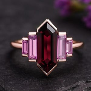 May include: A rose gold ring with a central, dark red gemstone and rectangular pink gemstones on either side. The ring has a geometric design. The background is dark, with purple flowers visible.