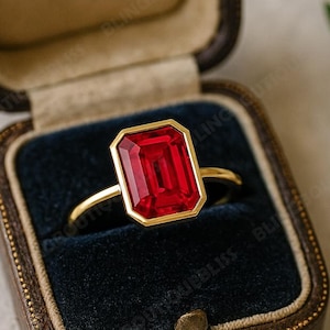 May include: A gold ring with a large, rectangular, red gemstone is displayed in a dark brown jewelry box with a blue velvet interior. The ring is the focal point, showcasing its vibrant color and elegant design. A sprig of lavender is in the background.