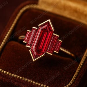 May include: A gold ring featuring a central rectangular red gemstone, with smaller red rectangular stones on either side. The ring is presented in a velvet jewellery box.