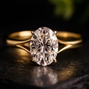 May include: A gold ring featuring a large, oval-cut diamond. The diamond is clear and faceted, reflecting light. The ring's band has a split design. The ring is set against a dark background.