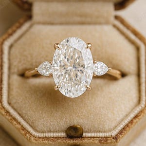 May include: A gold engagement ring with a large oval-cut diamond center stone and two pear-shaped side stones. The ring is set in a gold band and is displayed in a brown velvet ring box.