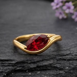 May include: A gold ring with a marquise-cut, deep red gemstone. The ring has a smooth, curved band and small gold accents on either side of the gemstone. The ring is displayed on a dark, textured surface.