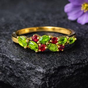 May include: A gold ring featuring a cluster of green and red gemstones. The ring's design incorporates oval-shaped green stones and smaller, round red stones, set in a gold band. The ring is displayed on a dark, textured surface.