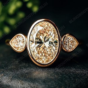 Könnte beinhalten: Ein goldener Ring mit einem großen, oval geschliffenen Diamanten als Mittelstein und zwei tropfenförmigen Seitensteinen. Die Diamanten haben einen warmen Champagnerton und sind in eine goldene Fassung gefasst. Der Ring wird auf einer dunklen, strukturierten Oberfläche präsentiert.