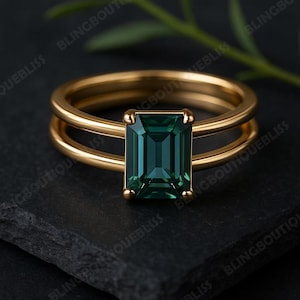 Può includere: Un anello d'oro con un design a doppia fascia. L'anello presenta una pietra preziosa di colore verde-blu, taglio smeraldo e forma rettangolare. L'anello è posto su uno sfondo scuro con un rametto di verde.
