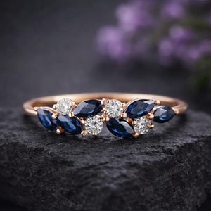 Puede incluir: Un anillo de oro rosa con un diseño floral con zafiros azules talla marquesa y diamantes blancos redondos. Las piedras preciosas están dispuestas en un delicado patrón en forma de hoja. El anillo se muestra sobre una superficie oscura y texturizada.