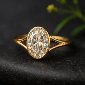 Könnte beinhalten: Ein goldener Ring mit einem oval geschliffenen Diamanten. Der Diamant ist in einer Lünettenfassung gefasst und das Band hat ein geteiltes Schaftdesign. Der Ring wird auf einer dunklen, strukturierten Oberfläche präsentiert.