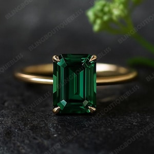 May include: A gold ring with a large, emerald-cut, deep green gemstone. The ring has a thin, gold band and four prongs holding the gemstone. The dark background accentuates the ring's features.