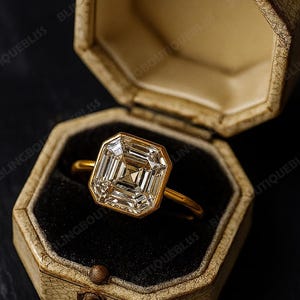May include: A gold ring with a large, square-cut diamond is displayed inside an open, vintage-style octagonal jewelry box. The ring's setting is gold, and the diamond is clear and multifaceted. The box is beige with a dark interior.