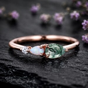 May include: A rose gold ring featuring a trio of stones: a small round clear gemstone, a pear-shaped iridescent opal, and a larger pear-shaped moss agate with green inclusions. The band is thin and polished, showcasing a delicate prong setting.