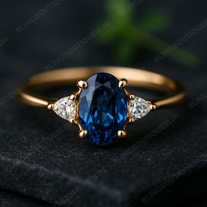 Könnte beinhalten: Ein goldener Ring mit einem großen, oval geschliffenen, tiefblauen Edelstein als Mittelpunkt, flankiert von zwei kleineren, birnenförmigen, klaren Edelsteinen. Der Ring liegt auf einer dunklen Oberfläche, die das Funkeln der Edelsteine hervorhebt.