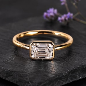 May include: A gold ring featuring an emerald-cut diamond. The ring has a slender, polished band, and the diamond is set within a bezel. The ring is presented on a dark surface, with purple flowers in the background. A piece of jewellery.