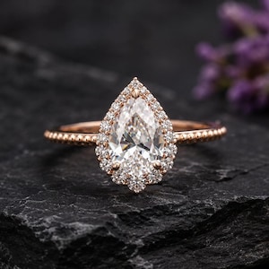 May include: A rose gold ring featuring a pear-shaped center stone surrounded by a halo of small round diamonds. The band has a delicate beaded texture along the sides, and the setting is crafted with prongs securing the central pear-cut gemstone.