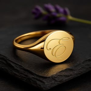 Pode incluir: Um anel de sinete dourado com um acabamento polido. O anel apresenta uma face circular gravada com a letra "E" em uma escrita elegante. O anel está sobre um fundo escuro, com um toque de flores roxas.