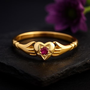 May include: A gold ring with a heart-shaped centerpiece and a red gemstone. The ring features stylized hands on either side of the heart. The ring is set against a dark background with a blurred purple flower.