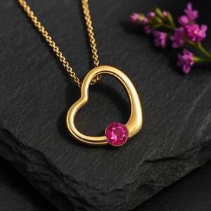 May include: A gold-colored heart-shaped pendant necklace with a bright pink gemstone set at the bottom. The necklace chain is also gold. The pendant is displayed on a dark, textured surface, with purple flowers in the background.