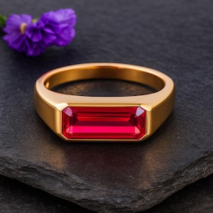 Könnte beinhalten: Ein goldfarbener Ring mit einem rechteckigen, leuchtend roten Edelstein. Der Ring liegt auf einer dunklen Oberfläche, im Hintergrund eine lila Blume. Das Design des Rings ist schlicht und elegant, geeignet für den Alltag oder besondere Anlässe.