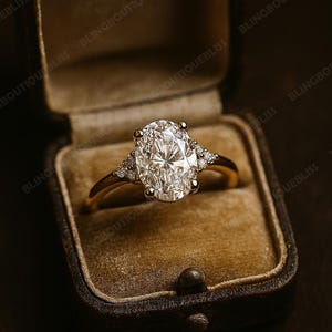 May include: A gold ring with a large oval-cut diamond center stone and smaller accent diamonds on each side. The ring is displayed in a vintage brown velvet ring box, creating a luxurious presentation.