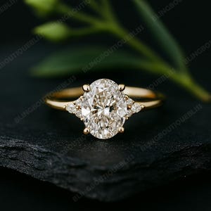 May include: A gold engagement ring with a large oval-cut diamond center stone and smaller accent diamonds on each side. The ring is set on a dark surface with a blurred green plant in the background.