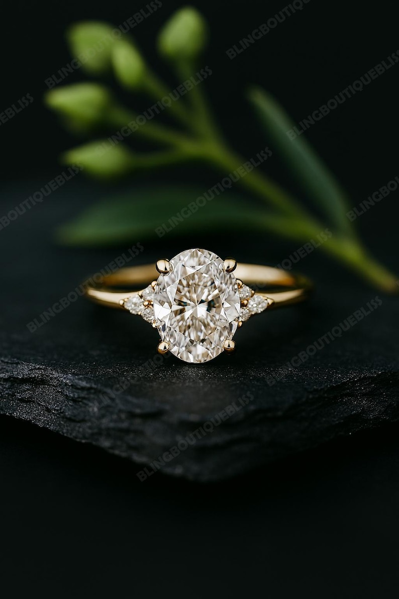 May include: A gold engagement ring with a large oval-cut diamond center stone and smaller accent diamonds on each side. The ring is set on a dark surface with a blurred green plant in the background.