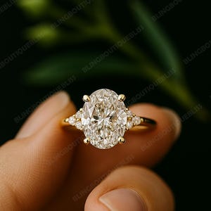 May include: A gold engagement ring with a large oval-cut diamond center stone and smaller accent stones on each side. The ring is held against a dark green background, highlighting the sparkle of the diamonds.