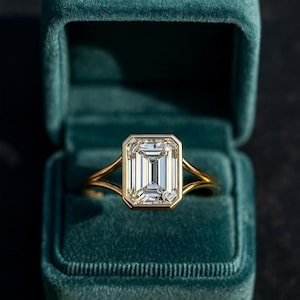 Może przedstawiać: Złoty pierścionek z dużym, prostokątnym diamentem o szlifie szmaragdowym w oprawie w kształcie ramki. Pierścionek jest prezentowany w otwartym, turkusowym aksamitnym pudełku na pierścionek. Obrączka ma rozszczepioną konstrukcję.