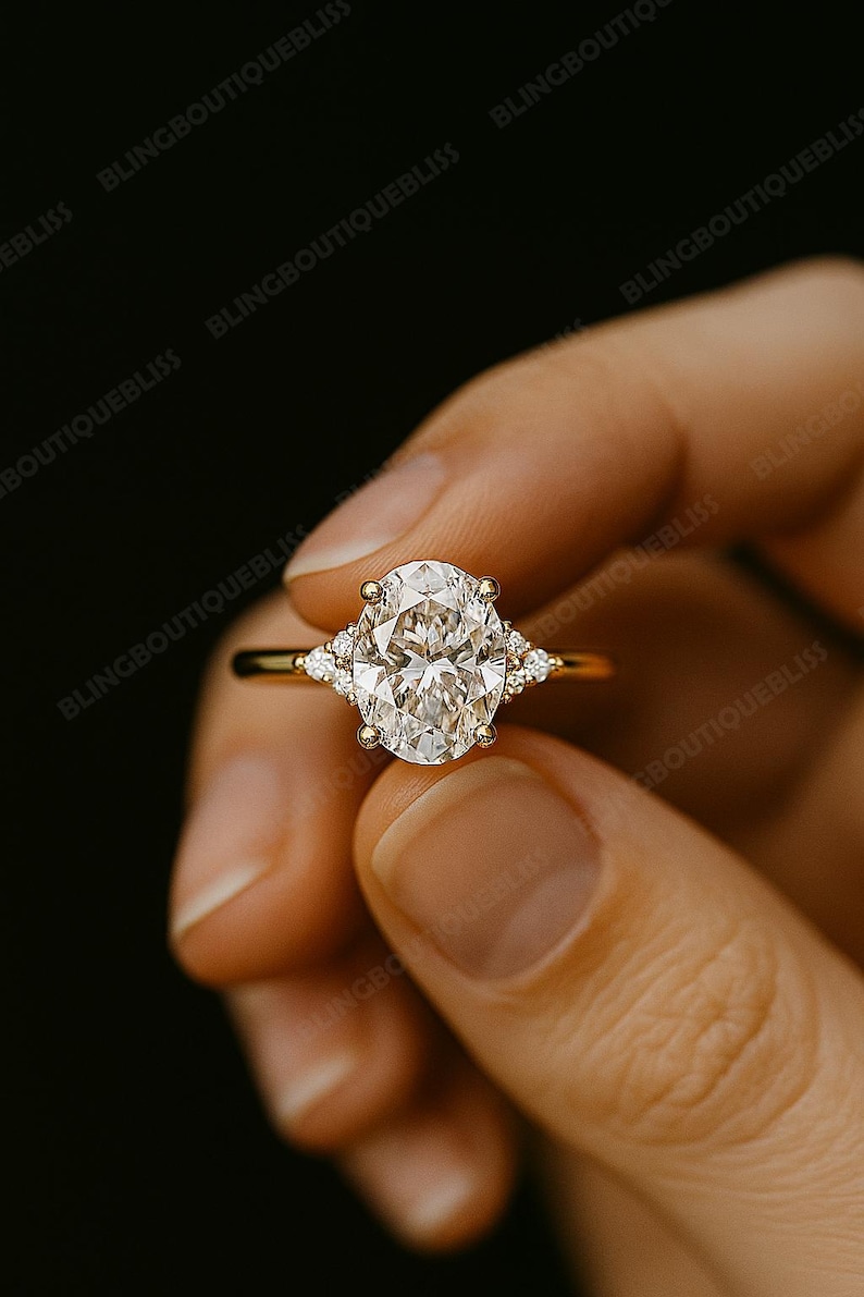 May include: A gold ring featuring a large oval-cut diamond center stone, flanked by smaller diamonds on each side. The ring is held against a dark background, highlighting the sparkle of the gemstones. The ring is a piece of fine jewelry.