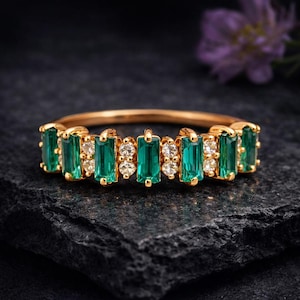 May include: A gold ring featuring alternating emerald-cut green gemstones and small, round, clear gemstones. The ring is set against a dark, textured surface, highlighting the sparkle of the gems. The ring is a piece of jewelry.