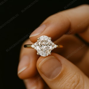 May include: A gold ring featuring a large oval-cut diamond center stone, flanked by smaller diamonds on each side. The ring has a delicate band and is held against a dark background. The ring is a piece of fine jewelry.