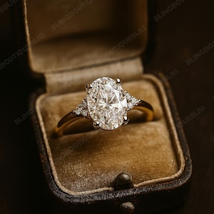 May include: A gold ring with a large oval-cut diamond center stone and smaller accent diamonds on each side. The ring is displayed in a vintage brown velvet ring box, creating a luxurious presentation.