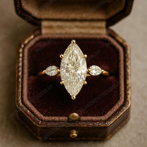 May include: A marquise-cut diamond ring with two smaller side stones, set in a gold band. The ring is displayed in a brown velvet ring box with a gold clasp. The ring's large center stone is the focal point.