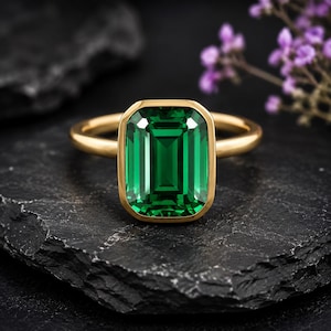 May include: A gold ring featuring a large, rectangular emerald-cut green gemstone set in a smooth bezel setting. The band is polished gold, and the center stone displays deep green facets that catch the light, creating a clear, vibrant appearance.