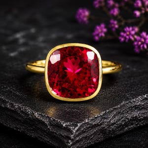 May include: A gold ring featuring a large, cushion-cut, vibrant red gemstone set in a smooth, polished bezel setting. The band is thin and circular, crafted from yellow gold, providing a clean and classic look that highlights the deep red colour.