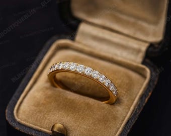 Può includere: Un anello a fascia in oro con una fila di pietre rotonde, chiare e sfaccettate lungo la metà superiore. L'anello è esposto all'interno di un portagioie rettangolare scuro, foderato in velluto, con coperchio incernierato e chiusura.