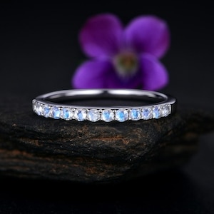 May include: A silver ring with a band of small, round, light blue gemstones. The ring is set against a dark background with a blurred purple flower in the background. The gemstones are evenly spaced around the band.
