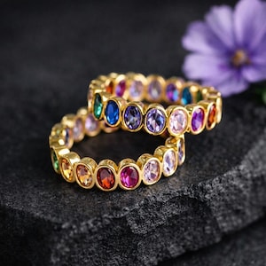 May include: Two gold-colored rings with a series of colorful, oval-shaped gemstones. The gemstones are in shades of red, orange, yellow, green, blue, and purple. The rings are displayed on a dark, textured surface.