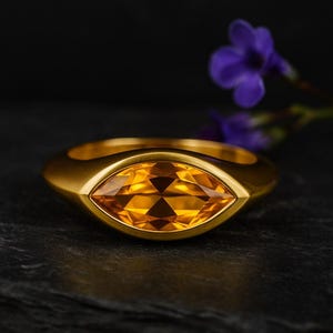 Peut inclure: Une bague en or avec une grosse pierre précieuse jaune-orangé facettée en forme d'œil. La bague a une finition lisse et mate. Une fleur violette est en arrière-plan.