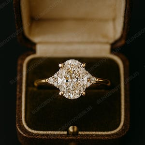 May include: A gold engagement ring with an oval-cut diamond center stone and smaller accent diamonds. The ring is set in a gold band and is displayed in a brown velvet ring box.