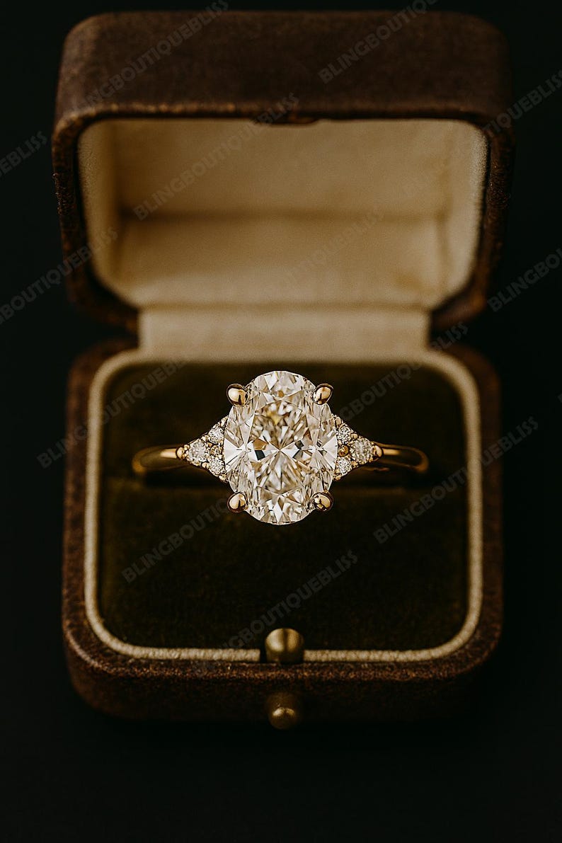 May include: A gold engagement ring with an oval-cut diamond center stone and smaller accent diamonds. The ring is set in a gold band and is displayed in a brown velvet ring box.