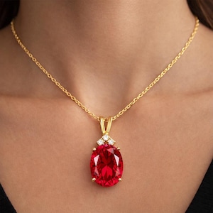 Peut inclure: Un collier doré avec un grand pendentif en pierre rouge de forme ovale. Le pendentif est orné de petits diamants transparents. La chaîne délicate est également dorée. Le collier est présenté sur le cou d'une personne.